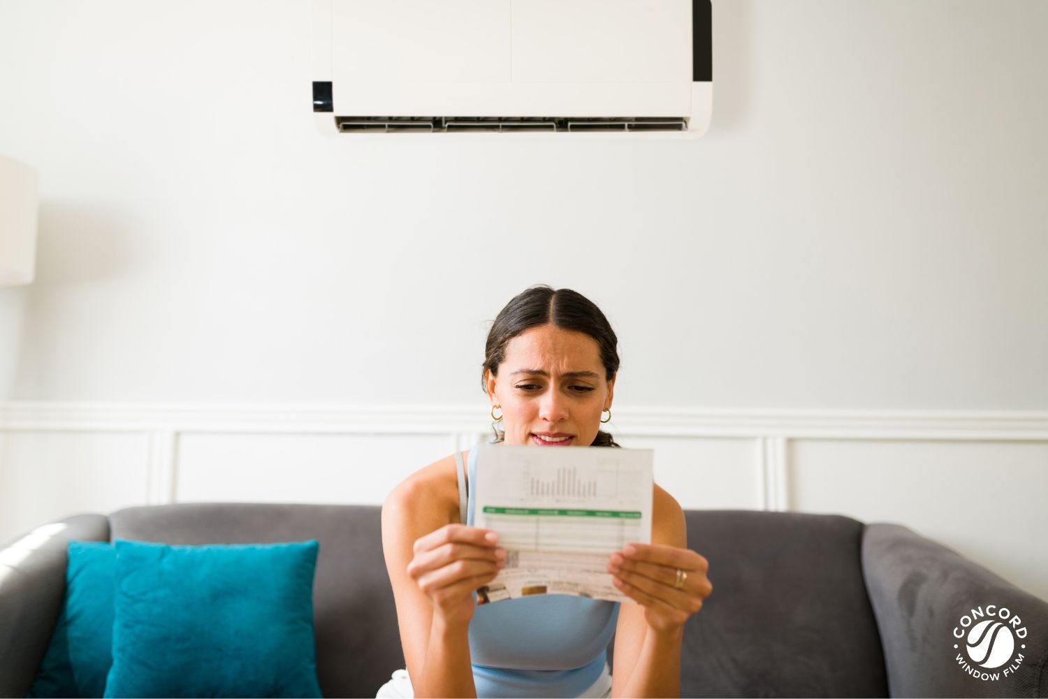 Woman sitting on the couch underneath an air conditioner holding an electricity bill and looking upset for a blog post entitled 2024 Summer Electricity Prices